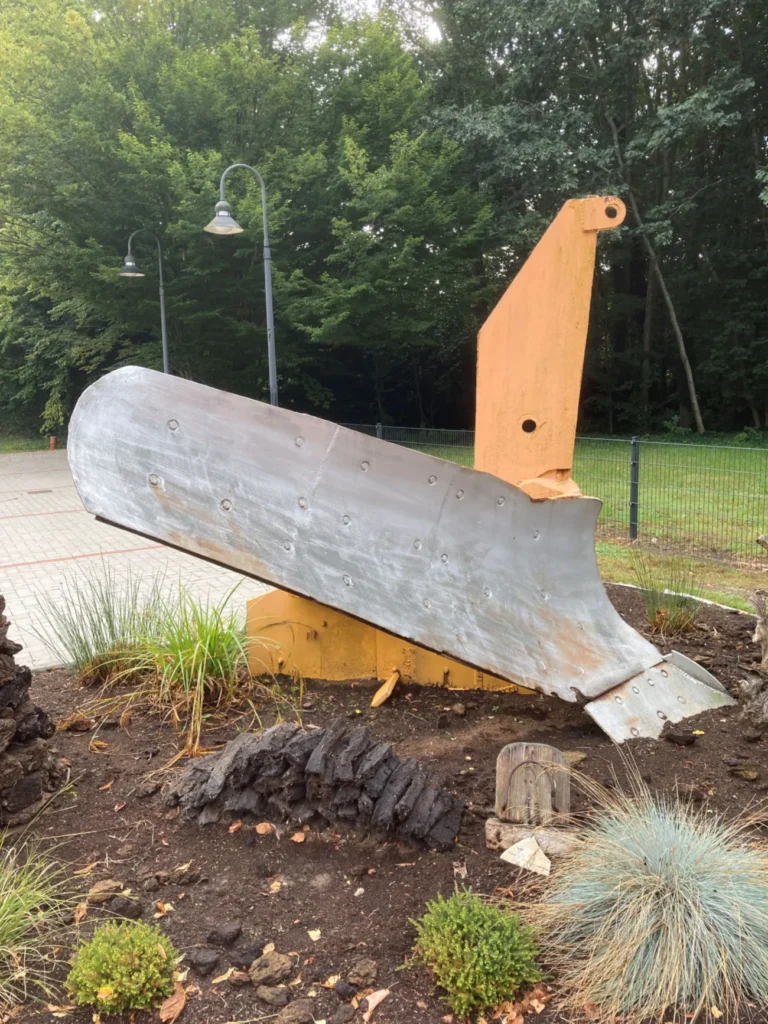 Gelber Baggerarm mit Schaufel als Freilicht-Denkmal in einem gepflegten Garten, Symbol der Moorerschließung.