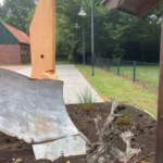 Rostiger Baggerarm mit Schaufel als Skulptur in einem Beet, daneben ein alter Baumstumpf und ein gepflasterter Weg.
