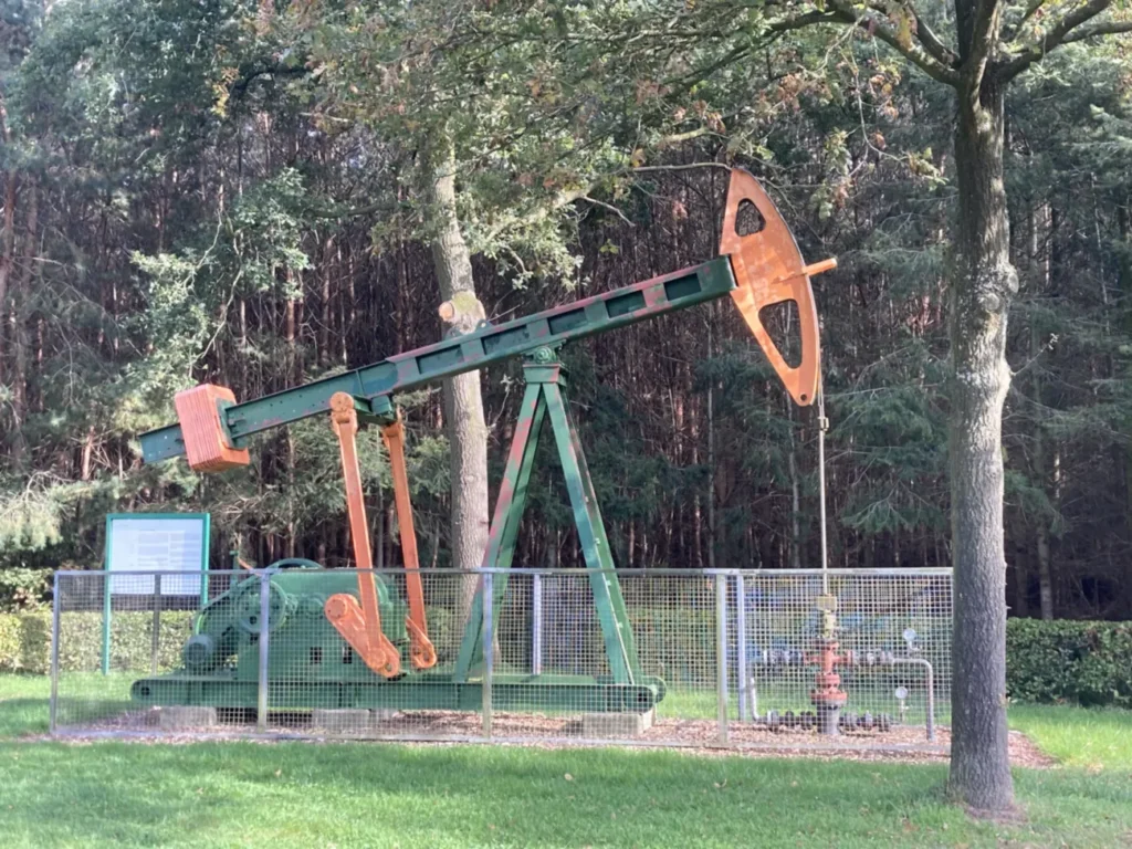 Erhaltene grün-orange Ölpumpe hinter Metallzaun in parkähnlicher Waldlandschaft als technisches Denkmal.