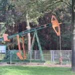 Erhaltene grün-orange Ölpumpe hinter Metallzaun in parkähnlicher Waldlandschaft als technisches Denkmal.
