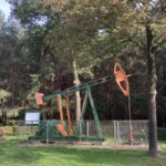 Restaurierte grün-orange Ölpumpe hinter Zaun mit Infotafel, umgeben von Bäumen, als technisches Denkmal im Emsland.