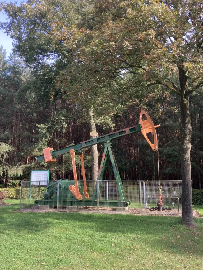 Restaurierte grün-orange Ölpumpe hinter Zaun mit Infotafel, umgeben von Bäumen, als technisches Denkmal im Emsland.
