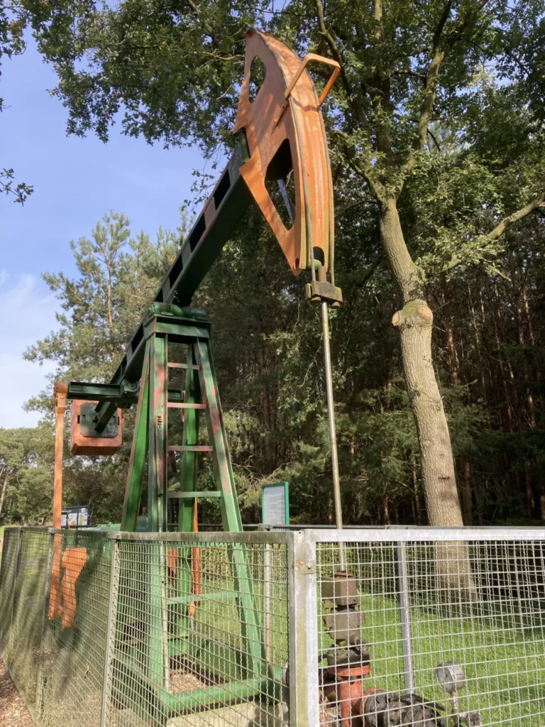 Erhaltene grün-orangefarbene Erdölpumpe hinter einem Metallgitterzaun, umgeben von Bäumen, als technisches Denkmal im Freigelände.