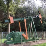 Historische Ölpumpe in Grün und Orange hinter einem Zaun, umgeben von Bäumen, als technisches Denkmal erhalten.