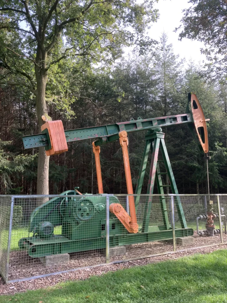 Historische Ölpumpe in Grün und Orange hinter einem Zaun, umgeben von Bäumen, als technisches Denkmal erhalten.