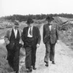 Drei Männer in Anzügen und Hüten spazieren auf einem Feldweg, umgeben von Heidelandschaft und Kiefernwald.