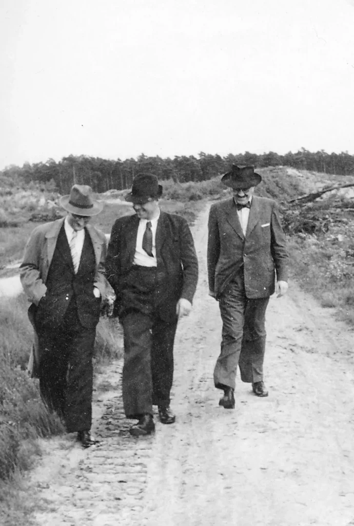 Drei Männer in Anzügen und Hüten spazieren auf einem Feldweg, umgeben von Heidelandschaft und Kiefernwald.