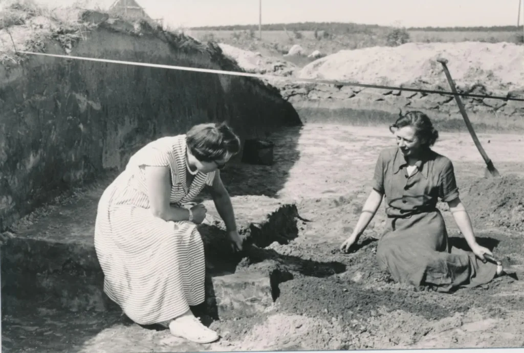 Zwei Frauen knien bei einer archäologischen Ausgrabung in einer Sandgrube und untersuchen Bodenfunde.