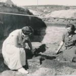 Zwei Frauen knien bei einer archäologischen Ausgrabung in einer Sandgrube und untersuchen Bodenfunde.