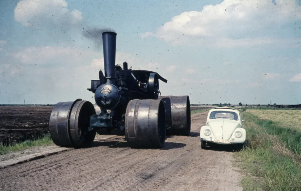 Eine riesige schwarze Ottomeyer-Lokomobile mit großen Stahlrädern fährt neben einem weißen VW Käfer auf einem Feldweg.