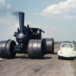 Eine riesige schwarze Ottomeyer-Lokomobile mit großen Stahlrädern fährt neben einem weißen VW Käfer auf einem Feldweg.