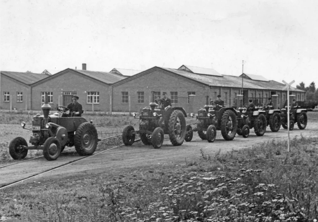 Sechs Traktoren mit Fahrern fahren in Kolonne an Backsteingebäuden vorbei, symbolisierend die frühe Motorisierung der Landwirtschaft.