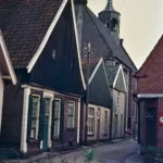 Blick in eine enge Kopfsteinpflasterstraße in Neuenhaus mit Backsteinhäusern und einem Kirchturm im Hintergrund.