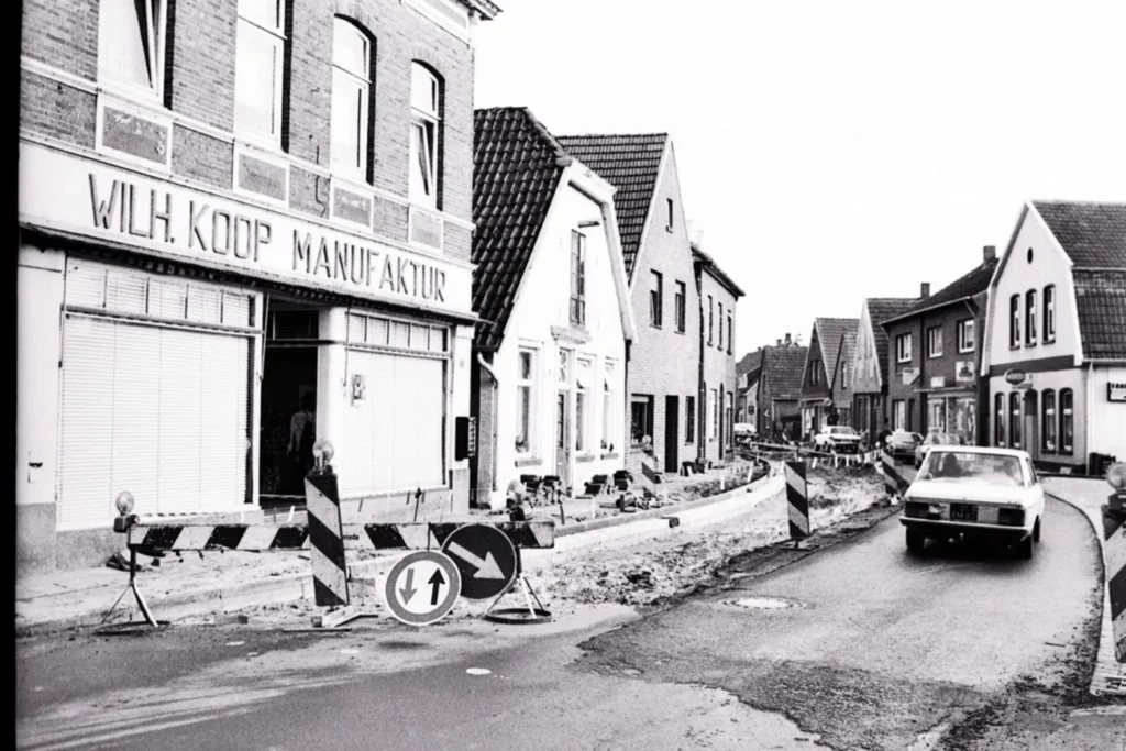 Schwarz-Weiß-Foto: Straßenbauarbeiten in Neuenhaus mit Absperrungen, vorbeifahrendem Auto und Geschäft Wilh. Koop Manufaktur.