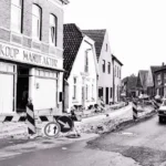 Schwarz-Weiß-Foto: Straßenbauarbeiten in Neuenhaus mit Absperrungen, vorbeifahrendem Auto und Geschäft Wilh. Koop Manufaktur.