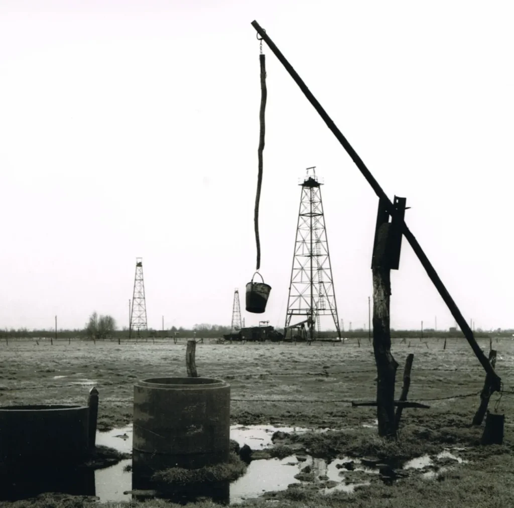 Schwarzweißfoto eines Ölfeldes mit mehreren Stahlfachwerk-Bohrtürmen, einem Brunnenkran mit Eimer und schlammigem Boden.