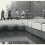 Gruppe von sieben Personen in formeller Kleidung besichtigt eine Kläranlage mit Betonbecken vor einem Backsteingebäude.