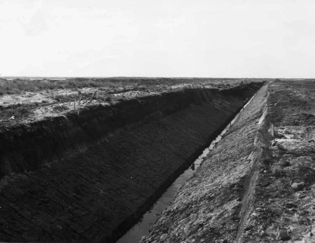 Schwarz-Weiß-Foto eines tief ausgehobenen Entwässerungskanals im Moor mit steilen Erdwänden und flachem Wassergrund.