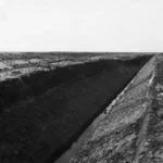 Schwarz-Weiß-Foto eines tief ausgehobenen Entwässerungskanals im Moor mit steilen Erdwänden und flachem Wassergrund.