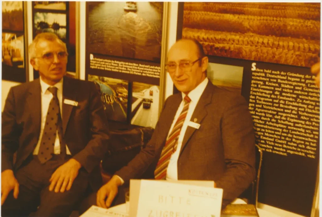 Zwei Männer in Anzügen sitzen an einem Messestand mit deutschen Informationstafeln im Hintergrund.