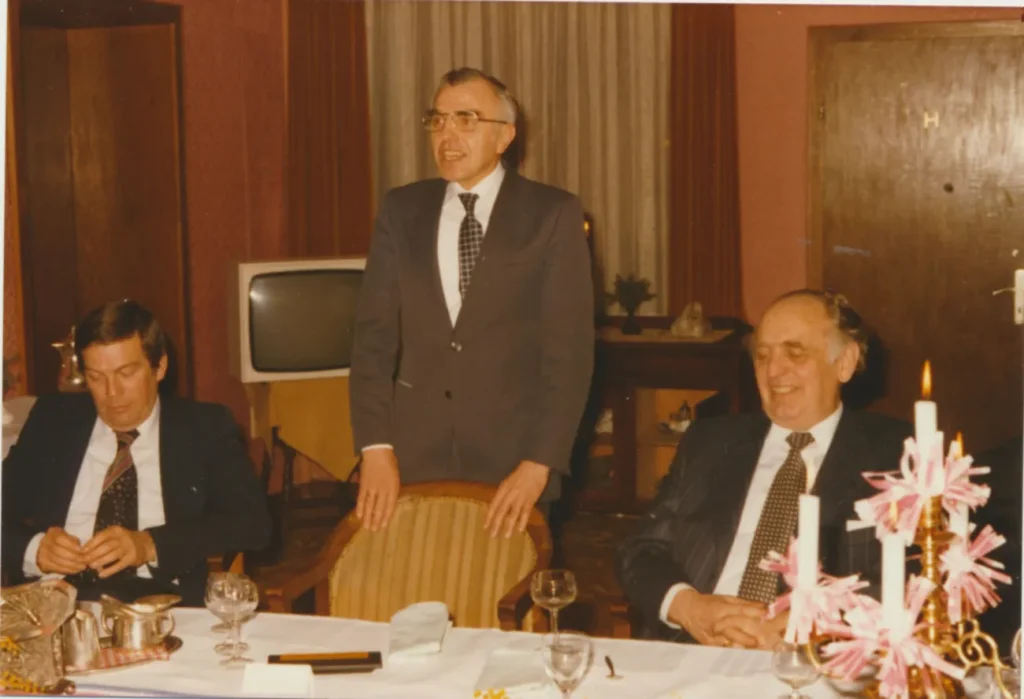 Ein stehender Mann in Anzug hält eine Rede bei einem festlichen Dinner, flankiert von zwei sitzenden Männern.