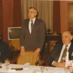 Ein stehender Mann in Anzug hält eine Rede bei einem festlichen Dinner, flankiert von zwei sitzenden Männern.