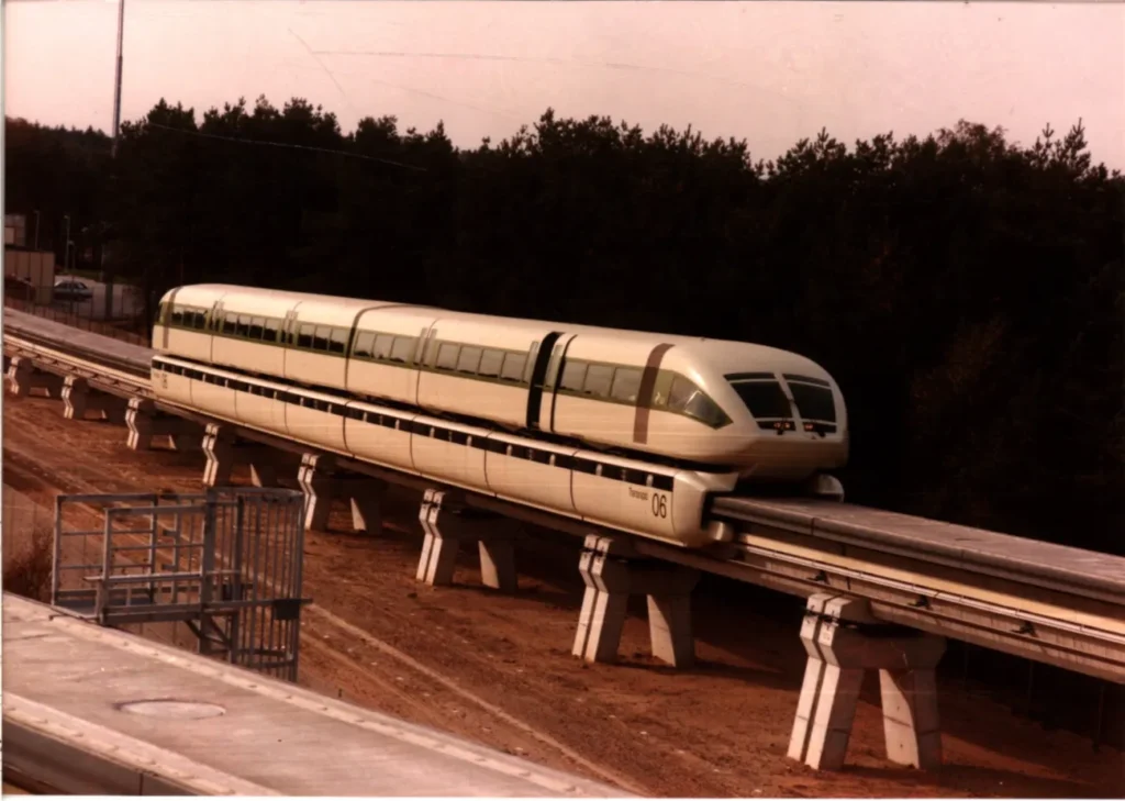 Der silberne Transrapid 06 schwebt auf einer erhöhten Betontrasse durch eine bewaldete Landschaft.