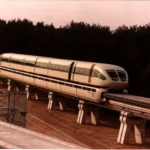 Der silberne Transrapid 06 schwebt auf einer erhöhten Betontrasse durch eine bewaldete Landschaft.