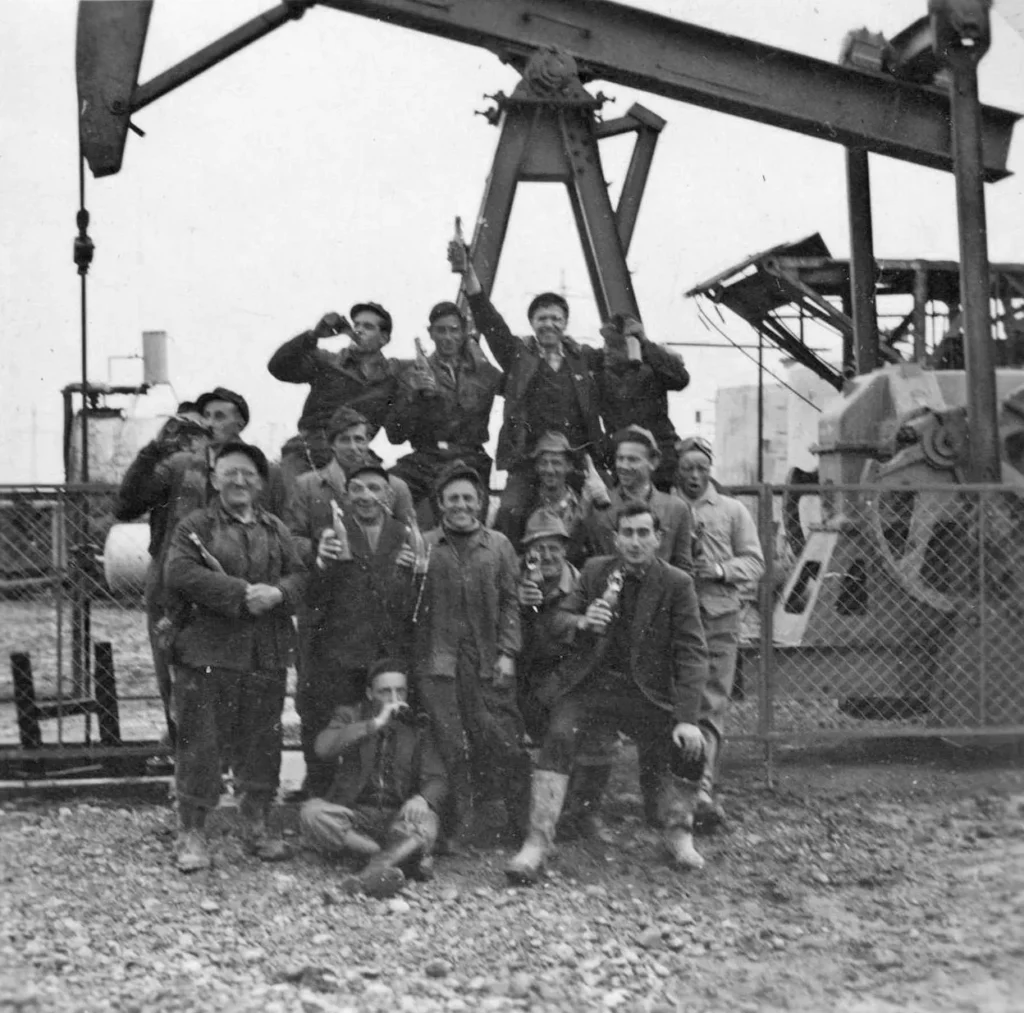 Gruppe von Arbeitern feiert mit Flaschen vor einer Erdölpumpe auf einem Schotterpatz, historische Schwarzweißaufnahme.