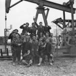 Gruppe von Arbeitern feiert mit Flaschen vor einer Erdölpumpe auf einem Schotterpatz, historische Schwarzweißaufnahme.