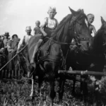 Zwei Kinder reiten auf Arbeitspferden beim Getreideschnitt, umgeben von Erwachsenen mit Erntewerkzeug.