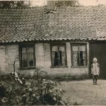 Historisches Foto eines einfachen Backsteinbauernhofs mit Ziegeldach, vor dem ein Kind und ein Hund stehen.