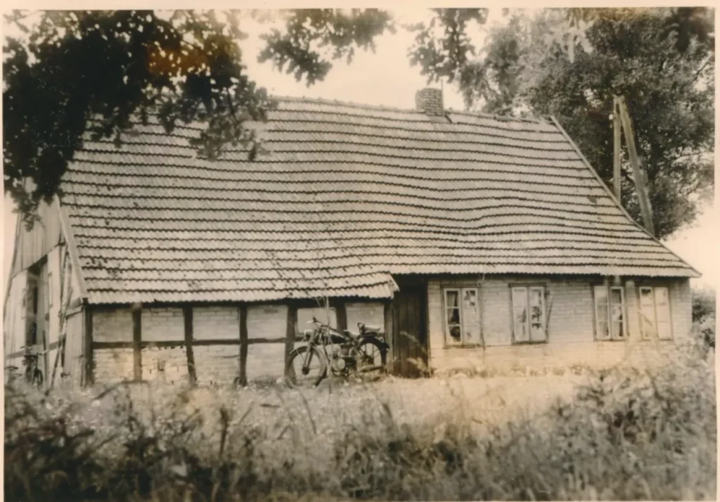 Altes, baufälliges Fachwerkgebäude mit Ziegeldach und einem Motorrad davor, umgeben von verwildertem Garten.