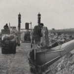 Historische Schwarzweißaufnahme: Zwei Traktoren ziehen einen Tiefpflug durch mooriges Gelände, begleitet von einem Arbeiter.