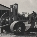 Zwei Arbeiter bedienen einen großen Tiefpflug vor einem Industriegebäude der Firma Gebrüder Kersten in Werlte.
