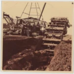 Historische Aufnahme einer Torfabbaumaschine mit Schaufelrad im Einsatz, zwei Arbeiter stehen daneben.