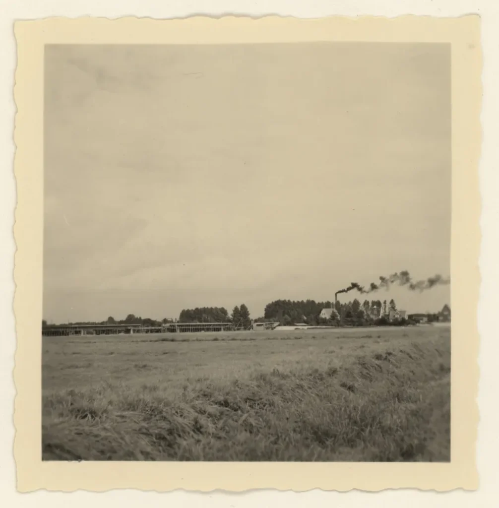 Schwarzweißfoto einer flachen Emslandlandschaft mit Fabrikgebäuden, rauchenden Schornsteinen und Bäumen am Horizont.