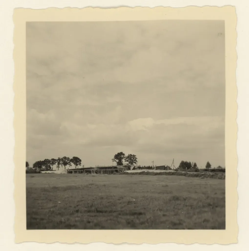 Historisches Schwarzweißfoto eines Gebäudes im Rohbau auf einer flachen Wiese mit Bäumen im Hintergrund.