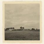 Historisches Schwarzweißfoto eines Gebäudes im Rohbau auf einer flachen Wiese mit Bäumen im Hintergrund.