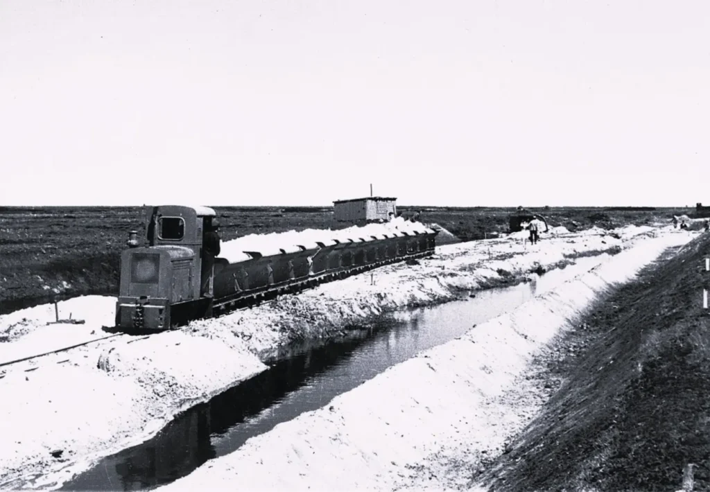 Eine Schmalspurbahn transportiert helles Material entlang eines Grabens durch flaches Moorland beim Straßenbau im Emsland.