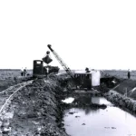 Ein Schaufelbagger gräbt einen Entwässerungskanal im Moor, begleitet von Arbeitern und einer Feldbahn.