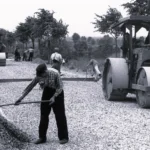 Arbeiter schaufeln Schotter, während zwei Dampfwalzen eine neue Straße im Emsland verdichten.