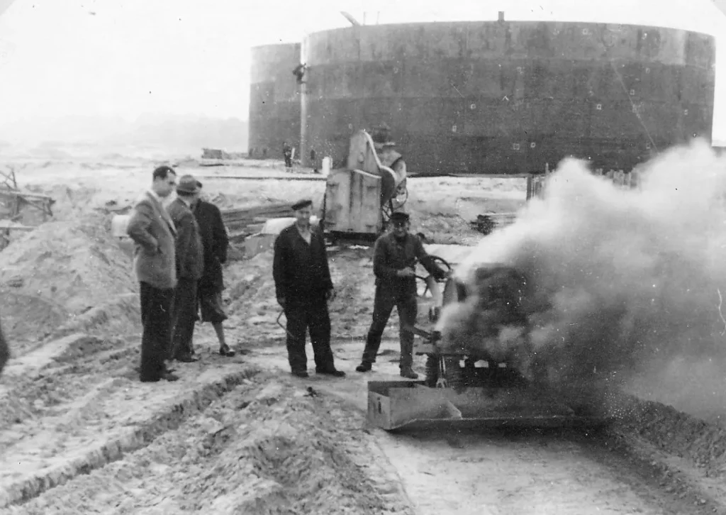 Arbeiter bedienen eine dampfende Maschine auf der Sandbaustelle der Raffinerie Lingen, im Hintergrund große Stahltanks.