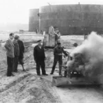 Arbeiter bedienen eine dampfende Maschine auf der Sandbaustelle der Raffinerie Lingen, im Hintergrund große Stahltanks.