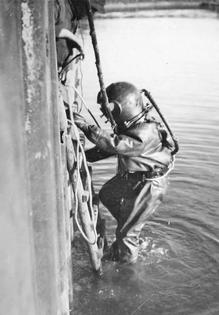 Ein Taucher in historischer Vollausrüstung arbeitet an einer Kanalwand im Wasser, vermutlich beim Bau der Raffinerie Lingen.