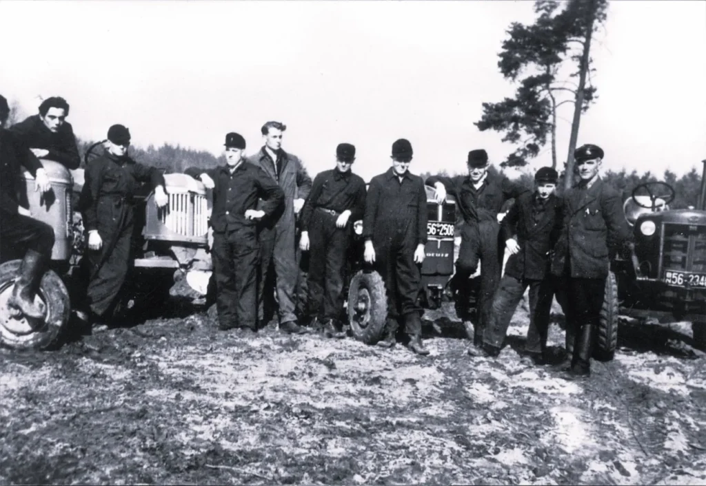 Gruppe von Männern in Arbeitskleidung posiert zwischen zwei Traktoren auf einem Feld, ca. 1950er Jahre.