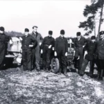Gruppe von Männern in Arbeitskleidung posiert zwischen zwei Traktoren auf einem Feld, ca. 1950er Jahre.