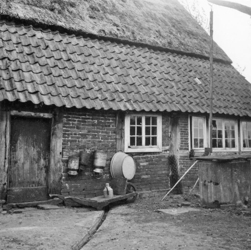 Historisches Schwarzweißfoto eines Bauernhauses mit Wassereimern und Handpumpe an der Außenwand, um 1950.