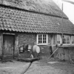 Historisches Schwarzweißfoto eines Bauernhauses mit Wassereimern und Handpumpe an der Außenwand, um 1950.