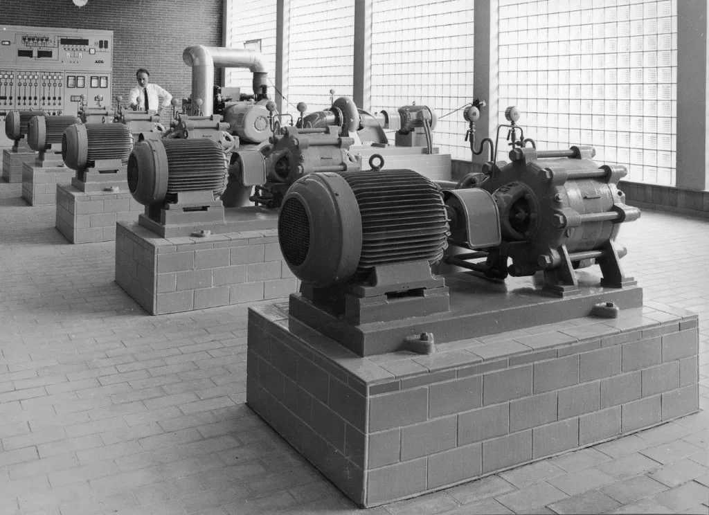 Maschinenraum eines Wasserwerks mit mehreren elektrischen Kreiselpumpen auf Betonsockeln und einem Bediener an der Schalttafel.